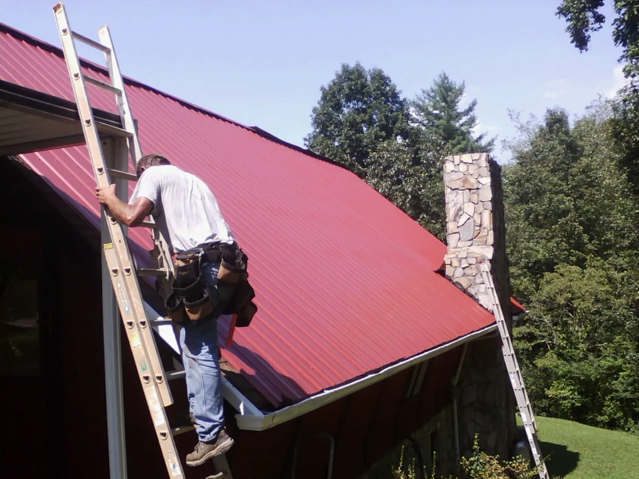 Roofer on a ladder beside a red metal roof and stone chimney — Soffit & Fascia Repair by Roofing Brothers in Mineral Wells